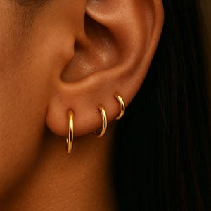 May include: Close-up of an ear adorned with three gold hoop earrings of varying sizes. The earrings are simple, circular, and shiny, reflecting light. The skin tone is warm, and the background is dark.