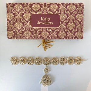 May include: A gold and white jeweled headpiece with a dangling pendant. The headpiece is in a box with the words "Kalo Jewelers" on it.