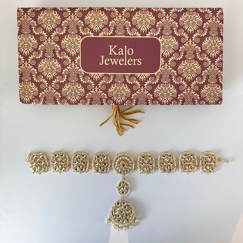 May include: A gold and white jeweled headpiece with a dangling pendant. The headpiece is in a box with the words "Kalo Jewelers" on it.