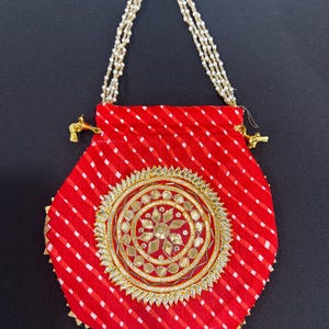 May include: A red fabric pouch with a white diagonal stripe pattern. The pouch features a gold and silver embroidered circular design in the center and a beaded handle. The pouch is approximately 6 inches wide.