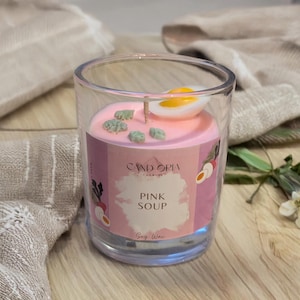 May include: A clear glass candle holder with a pink candle labelled "Pink Soup" by Candopia. The candle is decorated with small green stones and a slice of egg. The candle sits on a wooden surface.