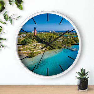 Puede incluir: Reloj de pared blanco con una vista aérea de un paisaje costero con un faro rojo, árboles verdes y agua turquesa. El reloj tiene manecillas y marcadores de hora negros y un marco blanco. Un artículo decorativo para cualquier habitación.