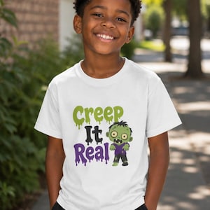Puede incluir: Una camiseta blanca de manga corta con un estampado gráfico que muestra el texto Creep It Real en letras verdes y moradas que gotean, acompañado de una ilustración de dibujos animados de un zombi verde con camisa morada y pantalones negros.