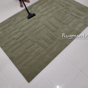 May include: A green patterned rug with a textured surface. The rug is being vacuumed by a person. The text "Rugmantra" is visible in the bottom right corner of the image.