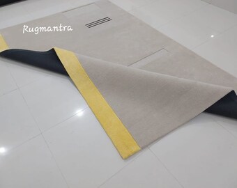 May include: A close-up of a beige rug with a yellow border and a black backing. The rug is folded in half, revealing the backing. The text "Rugmantra" is visible on the rug.