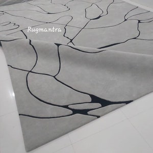 以下が含まれることがあります： 黒の抽象的なデザインが施された白い敷物。敷物は半分に折り畳まれており、両側にデザインが見えます。敷物は柔らかく、豪華な素材で作られています。画像の右上隅に「Rugmantra」というテキストが表示されています。
