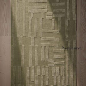 May include: A green and beige area rug with a geometric pattern. The rug has a textured surface and is made of wool. The rug is shown on a wooden floor. Rugmantra.