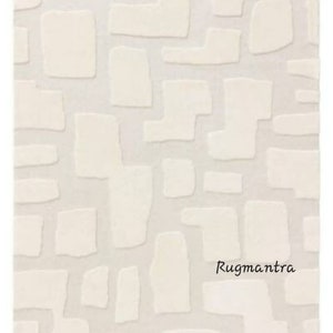 May include: Cream-colored rug with a geometric pattern of raised, irregular shapes. The shapes are in a lighter shade of cream, creating a subtle contrast against the background. The rug has a soft, textured appearance, suitable for home decor.