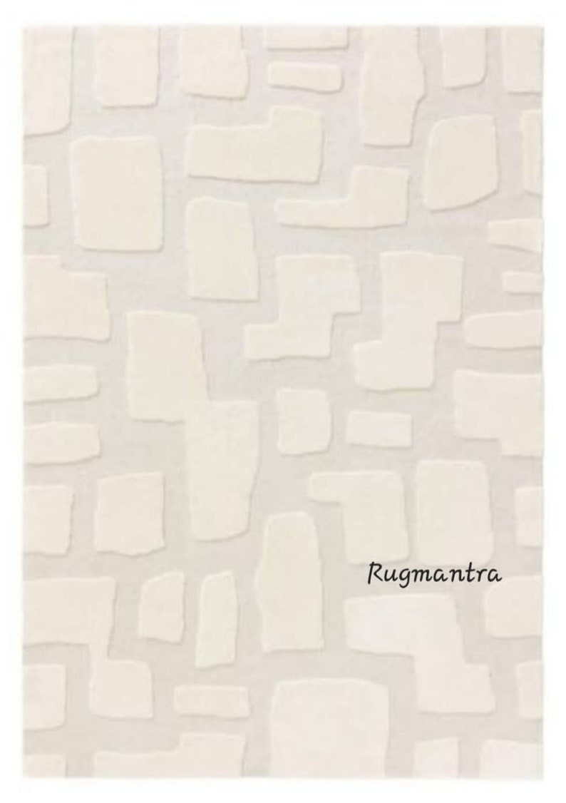 May include: Cream-colored rug with a geometric pattern of raised, irregular shapes. The shapes are in a lighter shade of cream, creating a subtle contrast against the background. The rug has a soft, textured appearance, suitable for home decor.