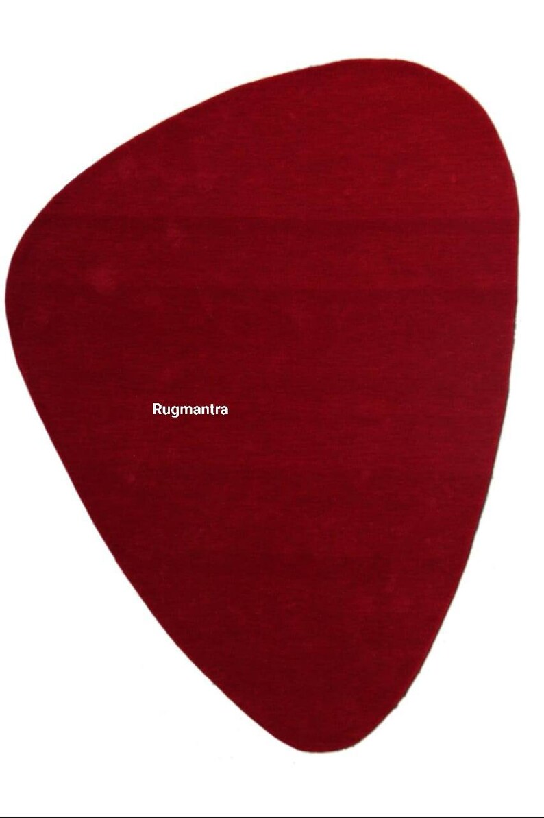 May include: A solid red, irregularly shaped rug with a slightly pointed bottom. The rug is made of a soft, plush material. The text "Rugmantra" is printed in white in the center of the rug.