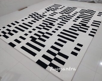 Può includere: Tappeto geometrico a motivi bianco e nero con sfondo bianco e strisce e quadrati neri. Il tappeto ha un design moderno ed è realizzato con materiali di alta qualità. Rugmantra è stampato nell'angolo in basso a destra.