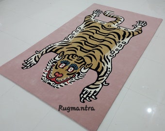 May include: A pink rug featuring a large, cartoon-style tiger print. The tiger is brown and black with white stripes and has a white face with black eyes and a red nose. The rug has the text "Rugmantra" printed in the bottom right corner.