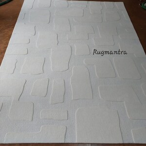 May include: A large, rectangular off-white rug with a textured, stone-like pattern. The design features raised, irregular shapes resembling cobblestones. The brand name "Rugmantra" is printed in black on the rug's surface. The rug is designed for home decor.