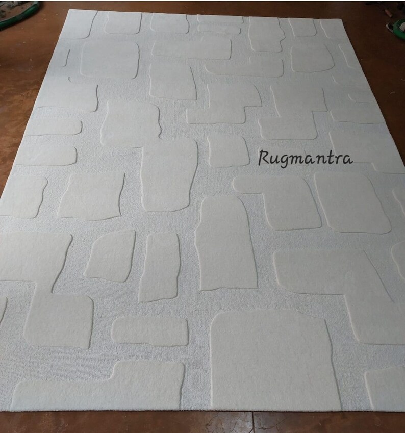 May include: A large, rectangular off-white rug with a textured, stone-like pattern. The design features raised, irregular shapes resembling cobblestones. The brand name "Rugmantra" is printed in black on the rug's surface. The rug is designed for home decor.