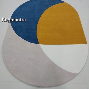 May include: A modern oval rug with geometric shapes in shades of blue, yellow, white, and pink. The rug features a large yellow circle with a blue crescent shape on top. The rug is made of soft, plush material.