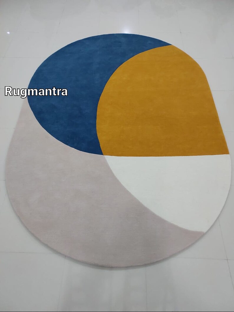 May include: A modern oval rug with geometric shapes in shades of blue, yellow, white, and pink. The rug features a large yellow circle with a blue crescent shape on top. The rug is made of soft, plush material.