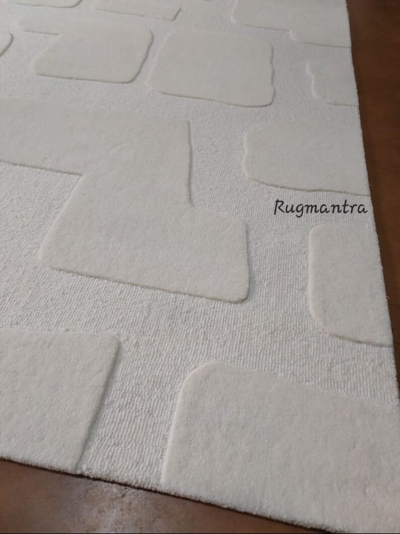 May include: Ivory-colored rug with a textured, stone-like pattern. The rug features raised, rectangular shapes in a lighter shade, creating a 3D effect. The design is set against a background of fine, vertical lines. The brand name "Rugmantra" is visible.