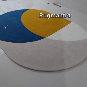 May include: A round, multi-colored rug with a blue, yellow, and white geometric design. The rug has a light beige background. The text "Rugmantra" is visible in the upper right corner of the image.
