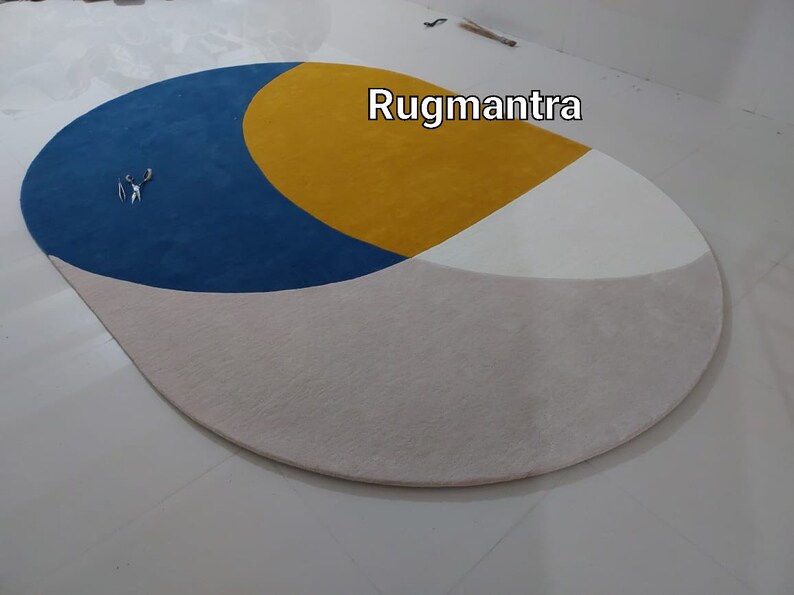 May include: A round, multi-colored rug with a blue, yellow, and white geometric design. The rug has a light beige background. The text "Rugmantra" is visible in the upper right corner of the image.