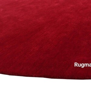 May include: A close-up of a deep red, plush rug with a soft, velvety texture. The rug is a solid color with no patterns or designs. The text "Rugmantra" is visible in the bottom right corner of the image.