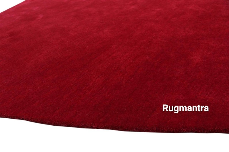 May include: A close-up of a deep red, plush rug with a soft, velvety texture. The rug is a solid color with no patterns or designs. The text "Rugmantra" is visible in the bottom right corner of the image.