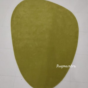 May include: A large, green, oval-shaped rug with a smooth texture. The rug is on a white tiled floor. The text "Rugmantra" is visible in the lower right corner of the rug.