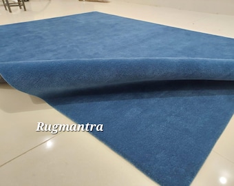 May include: A large, rectangular blue rug with a soft, textured surface. The rug is partially rolled up, revealing its underside. The text "Rugmantra" is visible in the lower left corner.