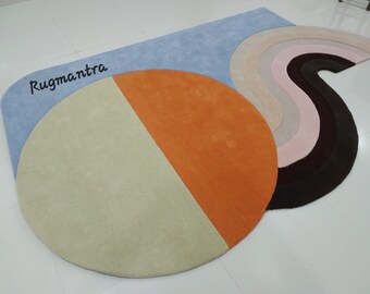 Puede incluir: Una colección de alfombras coloridas con diseños geométricos. La alfombra principal es azul claro con un gran círculo bicolor en beige y naranja. También se ve una alfombra curva con arcos de colores arcoíris y el texto "Rugmantra".