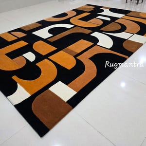 May include: Geometric patterned area rug in black, white, and shades of brown and orange. The rug features a repeating abstract design with curved and angular shapes. The text "Rugmantra" is visible in the lower right corner.