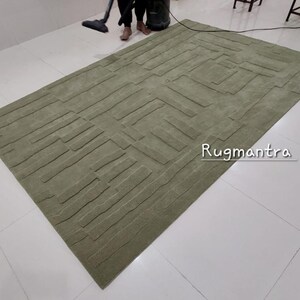 May include: A green area rug with a textured, geometric pattern. The rug has a modern design and is made of a soft, plush material. The text "Rugmantra" is visible in the lower right corner of the rug.