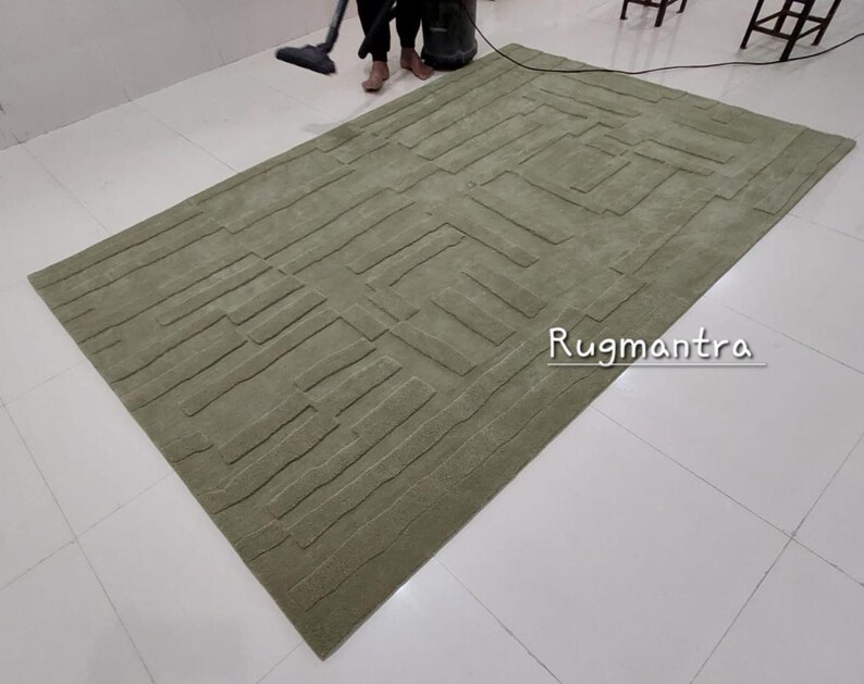 May include: A green area rug with a textured, geometric pattern. The rug has a modern design and is made of a soft, plush material. The text "Rugmantra" is visible in the lower right corner of the rug.