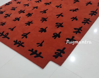 May include: A red rug with a black abstract pattern of leaves. The rug has a unique shape with a cut-out corner. The text "Rugmantra" is visible in the bottom right corner.
