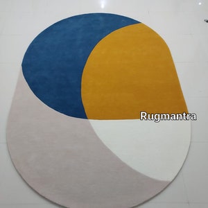 May include: A round, multi-colored rug with a blue, yellow, white, and pink geometric design. The rug has a slightly oval shape and the text "Rugmantra" is visible in the lower right corner.