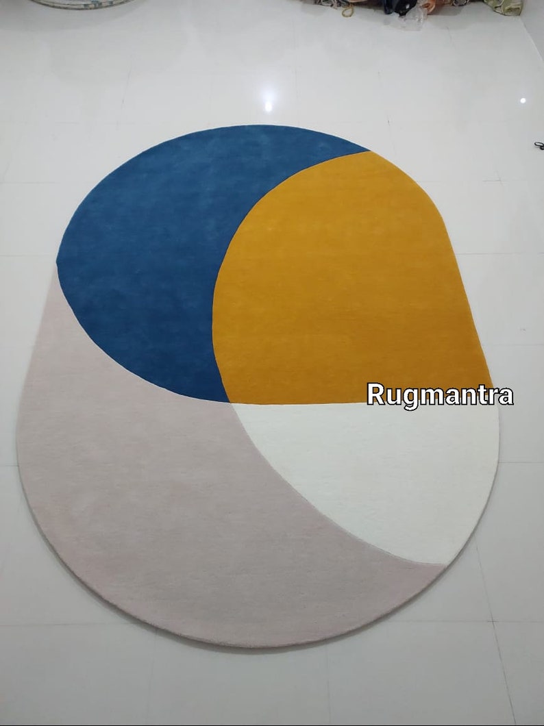 May include: A round, multi-colored rug with a blue, yellow, white, and pink geometric design. The rug has a slightly oval shape and the text "Rugmantra" is visible in the lower right corner.