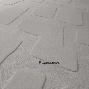May include: A white rug with a textured surface and a pattern of raised, irregular rectangular shapes. The rug's design features a soft, plush texture, creating a visually appealing and tactile experience. The text "Rugmantra" is visible on the rug.