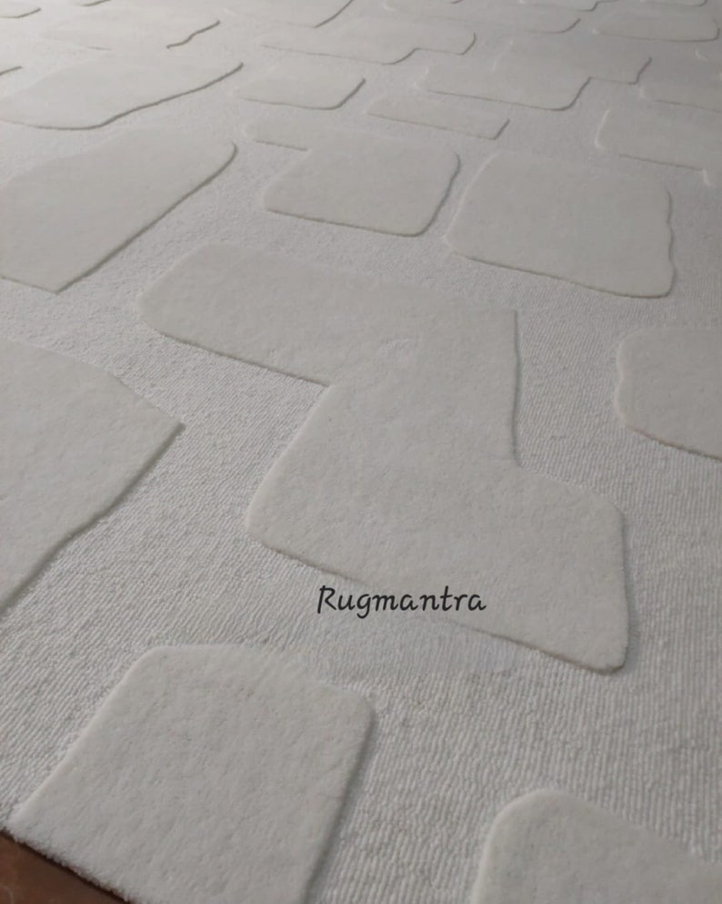 May include: A white rug with a textured surface and a pattern of raised, irregular rectangular shapes. The rug's design features a soft, plush texture, creating a visually appealing and tactile experience. The text "Rugmantra" is visible on the rug.