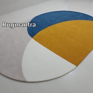 May include: A round rug with a geometric design featuring three colors: pink, white, and yellow. The yellow section is a half-circle with a blue half-circle on top. The text "Rugmantra" is visible in the upper left corner of the image.