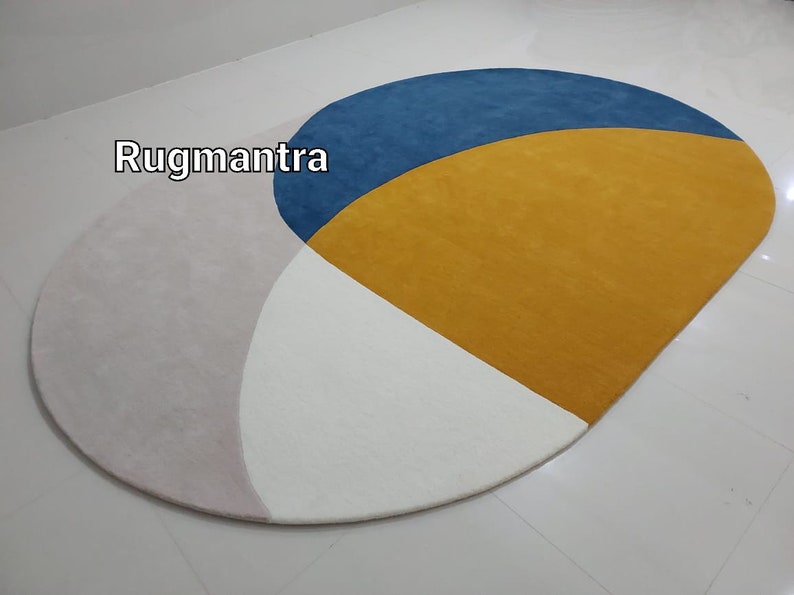 May include: A round rug with a geometric design featuring three colors: pink, white, and yellow. The yellow section is a half-circle with a blue half-circle on top. The text "Rugmantra" is visible in the upper left corner of the image.