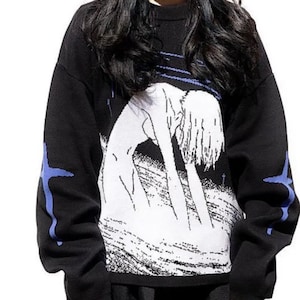 May include: Black sweater with a white graphic print of a woman with long hair. The sweater has blue accents on the sleeves.