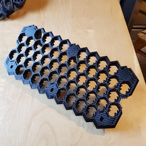 May include: A black 3D printed grid organiser featuring a repeating hexagonal pattern with textured carbon fibre-like surfaces on the edges. The structure consists of multiple interconnected hexagonal cells designed for storage and organisation.