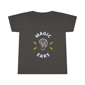 Peut inclure: T-shirt gris foncé pour tout-petits avec les mots "MAGIC EARS" en blanc, avec un dessin d'oreille blanche avec une aide auditive et des éclairs jaunes. Le t-shirt est en matière souple.