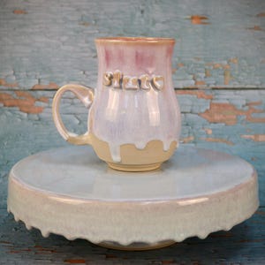 Pode incluir: Uma caneca de cerâmica com acabamento em esmalte brilhante rosa e branco, apresentando a palavra "sluts" em letras texturizadas em relevo na frente. A caneca repousa sobre um suporte circular de cerâmica correspondente com esmalte azul claro e branco.
