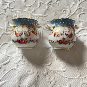 May include: Two white porcelain urns with blue and gold accents and floral designs. The urns have two handles on each side and a scalloped rim.