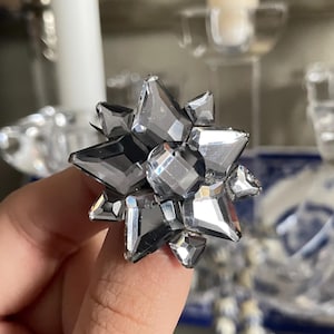 Pode incluir: Um anel prateado em forma de estrela com múltiplas facetas. O anel é feito de um material refletor, que capta a luz e cria um efeito brilhante. O anel é segurado pelo dedo de uma pessoa.