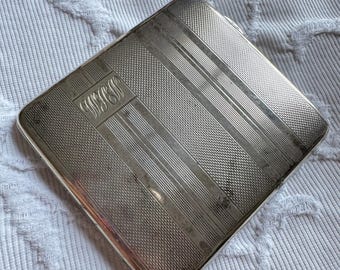 May include: A silver cigarette case with a monogrammed design and a textured surface.
