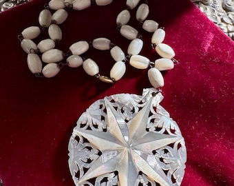Puede incluir: Un collar vintage con un colgante en forma de estrella y cuentas ovaladas. El colgante está intrincadamente tallado. El collar se exhibe sobre una superficie de terciopelo rojo. Las cuentas y el colgante son de color nacarado.