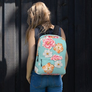 Può includere: Uno zaino azzurro chiaro con un motivo floreale. I fiori sono rosa, arancioni e bianchi. Lo zaino ha una tasca con cerniera sul davanti.