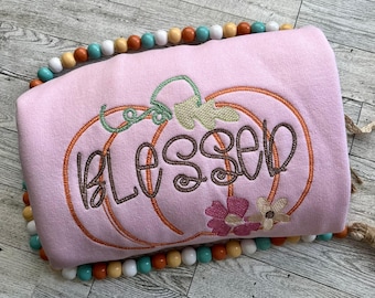 May include: A pale pink sweatshirt featuring an embroidered pumpkin design. The pumpkin outline is orange, with green vines and the word "Blessed" in brown script. Small pink and white flowers are also embroidered.