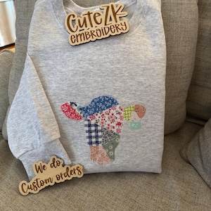 Puede incluir: Sudadera gris claro con un diseño de vaca de retazos. La cabeza de la vaca está hecha de varias telas estampadas y florales. La sudadera tiene cuello redondo y mangas largas. Las etiquetas de madera dicen "Cute ZK Embroidery" y "We do custom orders."