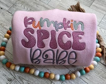 May include: A pale pink sweatshirt embroidered with the words "Pumpkin Spice Babe" in multiple colours. A beaded garland in orange, teal, and white surrounds the sweatshirt. A decorative tassel is on the right side.